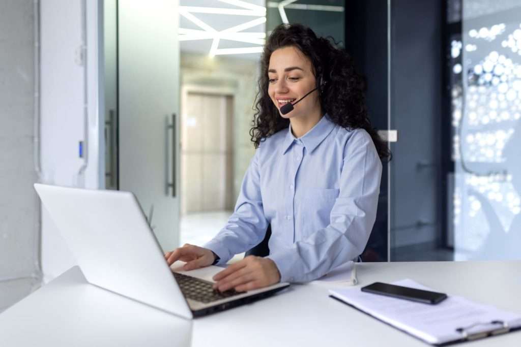 Smiling woman wearing a headset working on a laptop in a modern office, representing the responsive support offered by IT managed services providers.